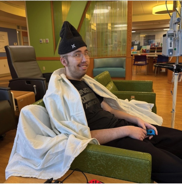 Alex wrapped in a blanket in a hospital treatment chair, smiling and holding a handheld game console